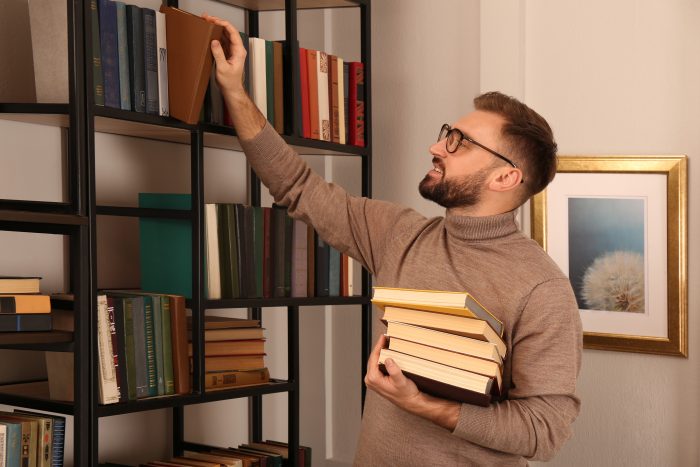 Man in a turtleneck holding a stack of books as he organizes a bookshelf.